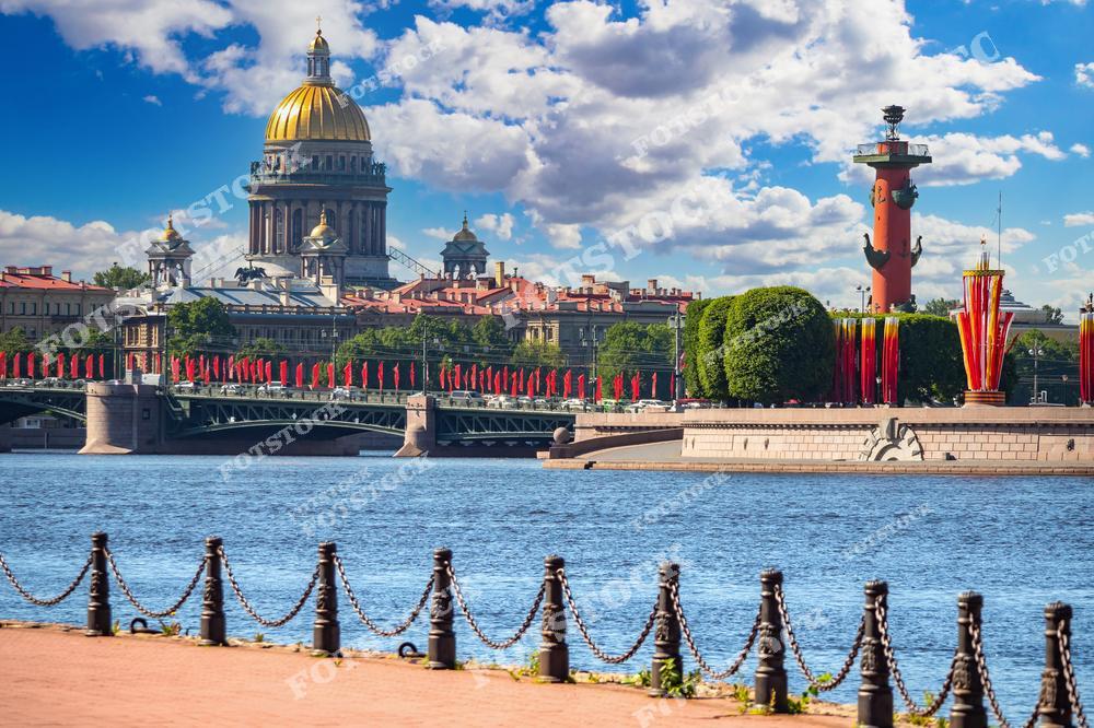 На фотографии изображены знаковые достопримечательности Санкт-Петербурга — Исаакиевский собор и Ростральные колонны на Стрелке Васильевского острова. Красные флаги и декоративные элементы на набережной создают торжественную атмосферу, указывая на праздничное событие. Собор с золотым куполом величественно возвышается на фоне яркого неба. Вид на Неву и городской пейзаж подчёркивает величие и красоту исторического центра города.